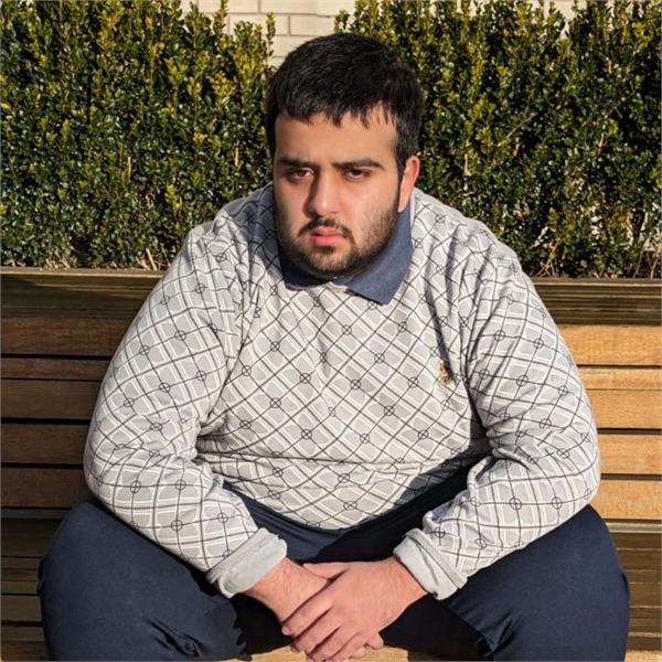 Hamza has black hair and a beard and is sitting down wearing a pattern jumper with blue trousers