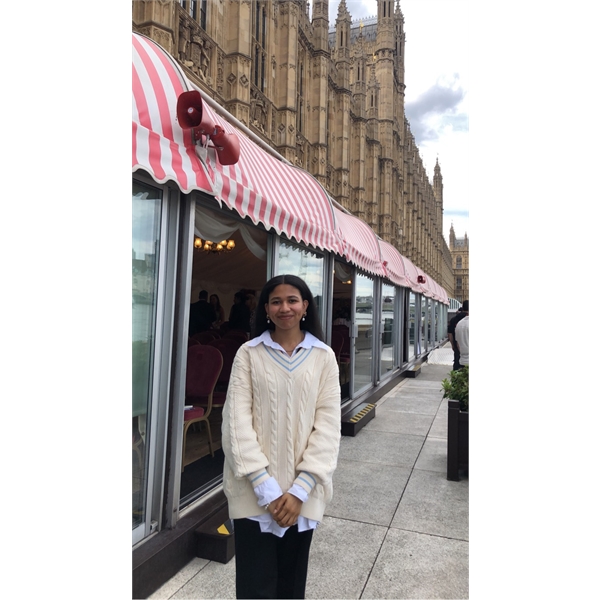 Khadija has black hair and is standing outside of a shop holding her hands together wearing a long white jumper with light blue shirt underneath and black trousers