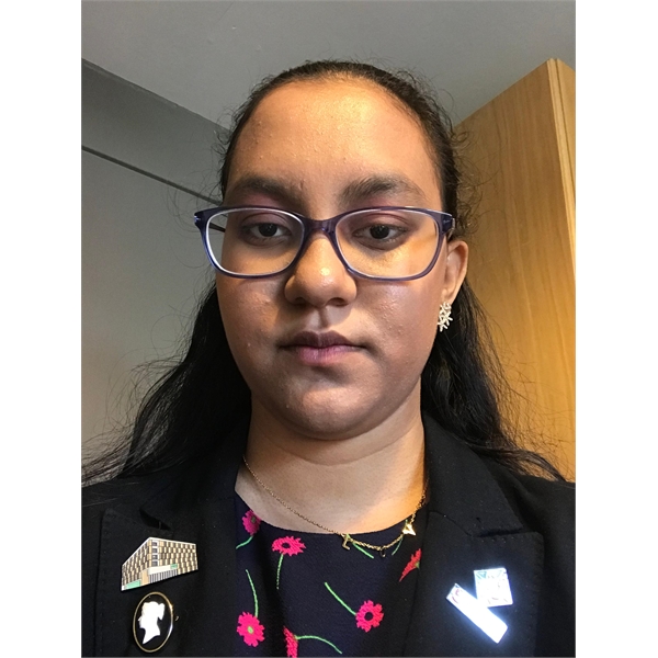 Rhea has long black hair and is wearing glasses with a rose pattern top and black shirt over the top of it