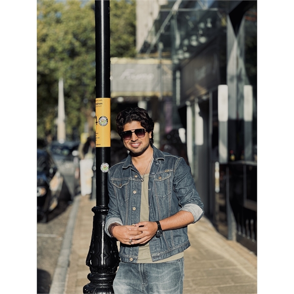 Sadais has long black hair and is wearing sunglasses whilst standing in front of a lamppost holding his hands together. He is wearing a denim shirt with rolled up sleeves and jeans