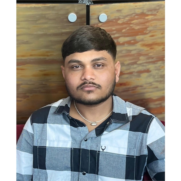 Sahil has black hair and a beard and sitting down and wearing a black and white chequered shit