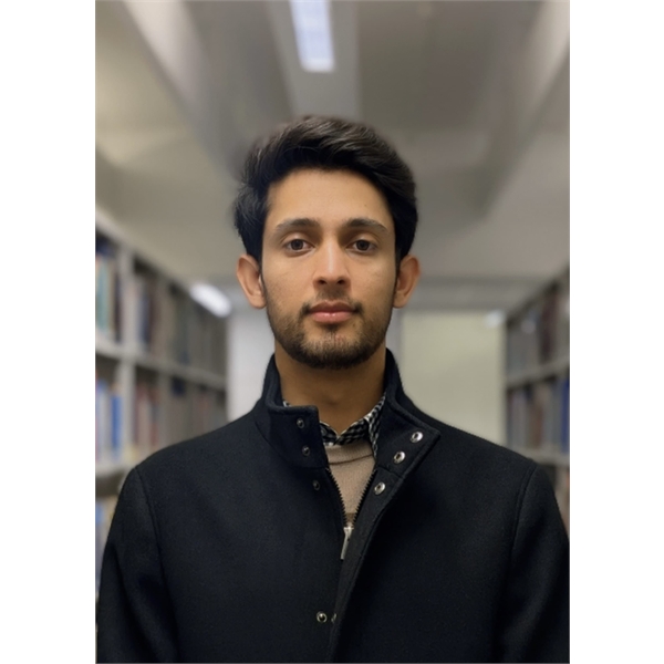 Syed has black hair and a beard and is wearing a black coat