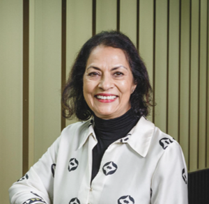 Anita is standing wearing a white loose flowy top with designs on with a long black top underneath and she has brown hair with red lipstick on