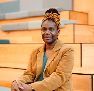 Imgoeen is sitting down in a study hall, wearing a beige coat wih a green top and black trousers whilst wearing an orange headband.