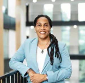 Marcia is standing on a stairwell smiling at the camera wearing a sky blue blazer and trousers with a white top underneath. Marcia has black hair and is wearing a blue necklace