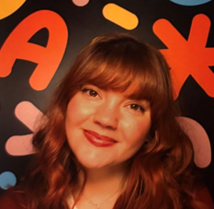 Rosemarie has red hair and red lipstick and smiling at the camera. She is wearing a stripy top and red cardigan