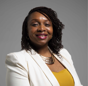 Yemisi has black hair with red lipstick. Yemisi is standing with her hands crossed at here front, wearing a silver jewllery and a white blazer with an orange top.